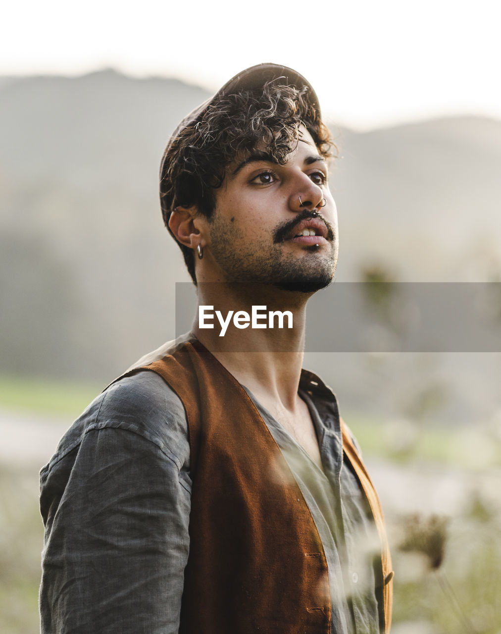 Dreamy young curly haired bearded ethnic male with piercing in ear wearing stylish clothes and cap looking away while standing on green meadow against blurred mountains