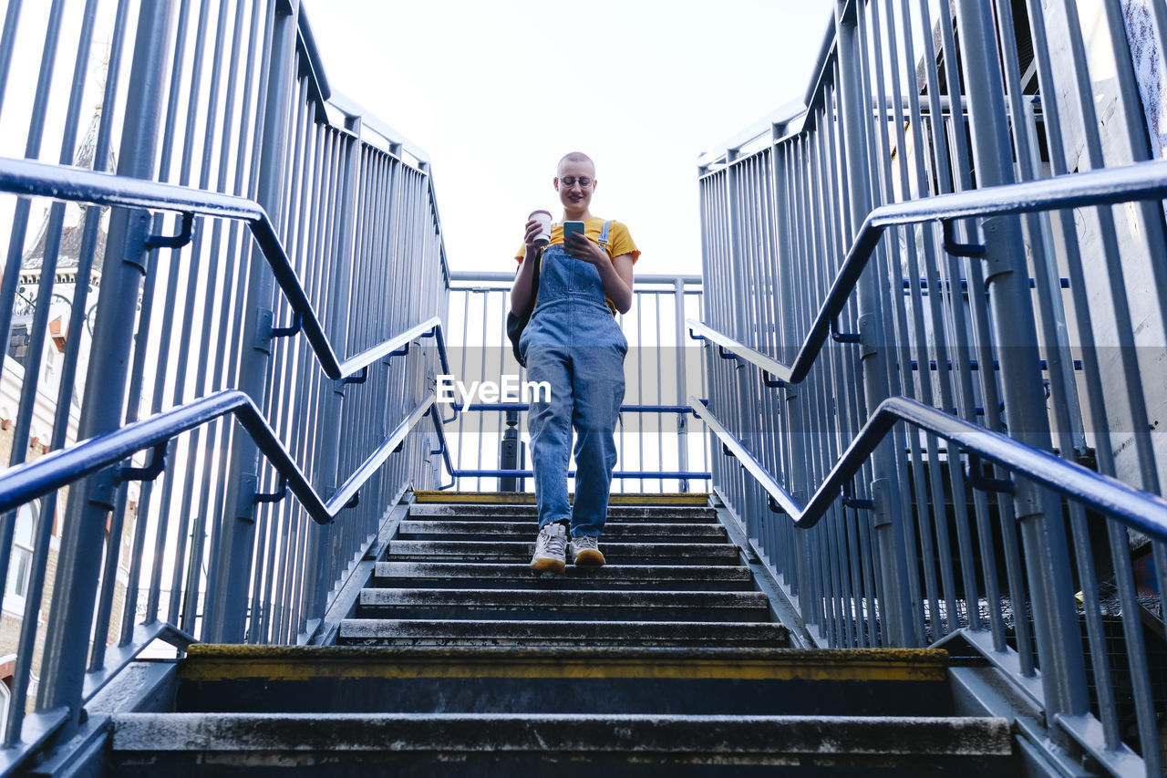 Non-binary person with disposable coffee cup using smart phone on steps