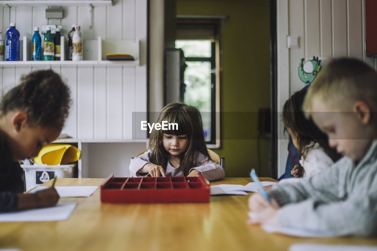 Multiracial male and female students writing on paper at table in kindergarten