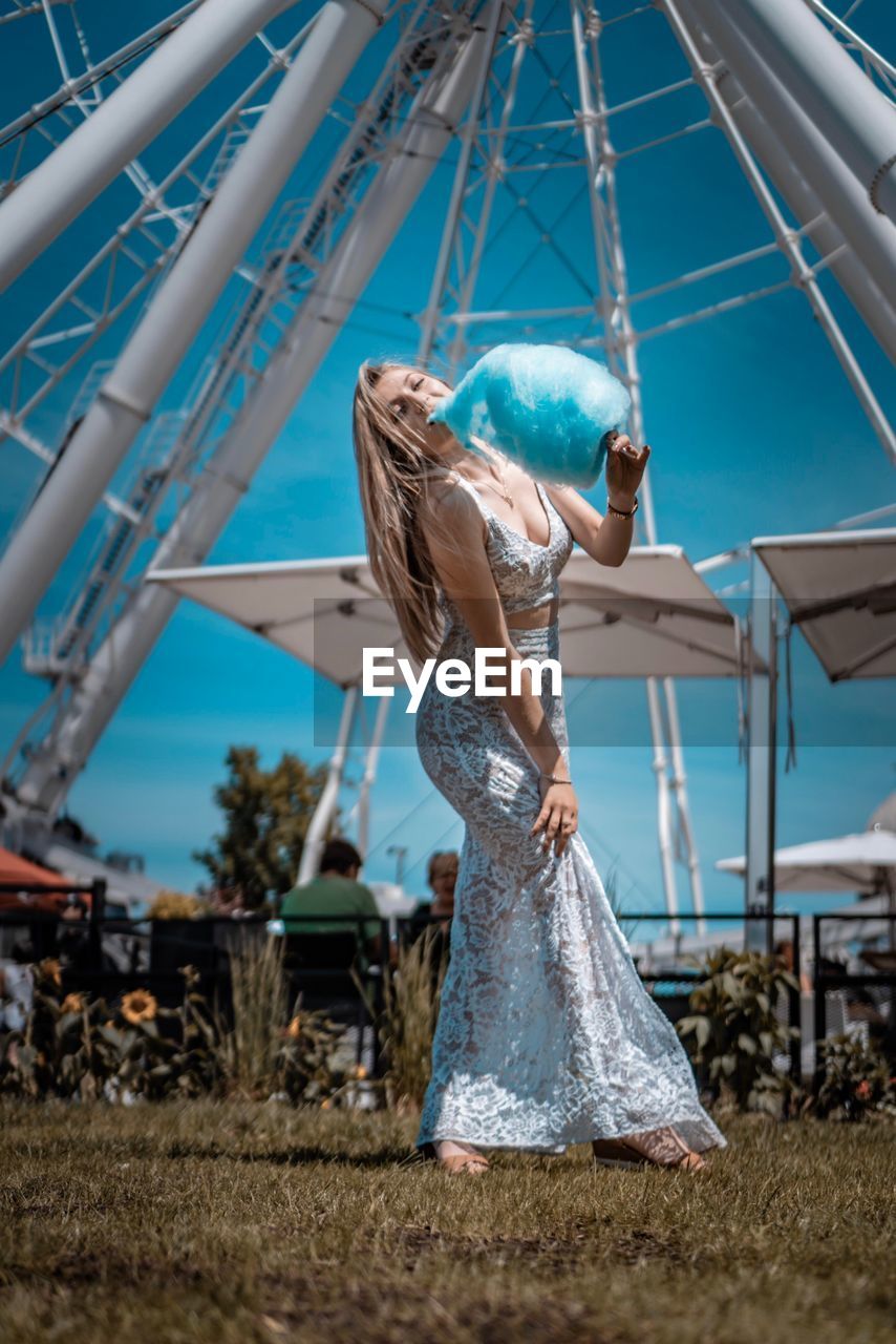 Full length of young woman holding cotton candy against ferris wheel on land