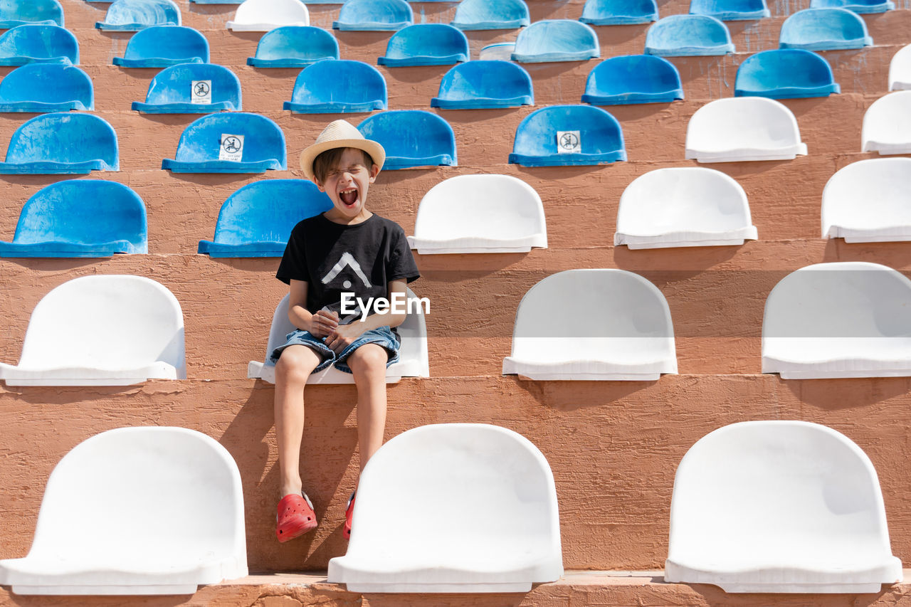 Boy in summer clothes sitting on one of chair on stadium and shout out with close eyes