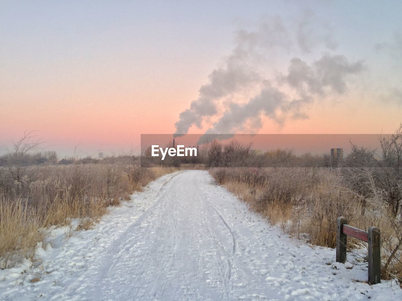 Snow covered pathway leading towards industry against sky during sunset