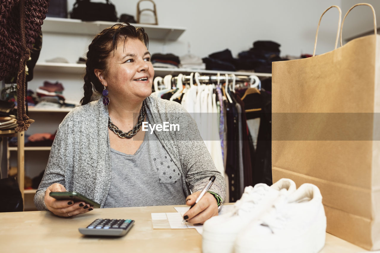 Smiling cashier counting with smart phone on checkout table in clothing store