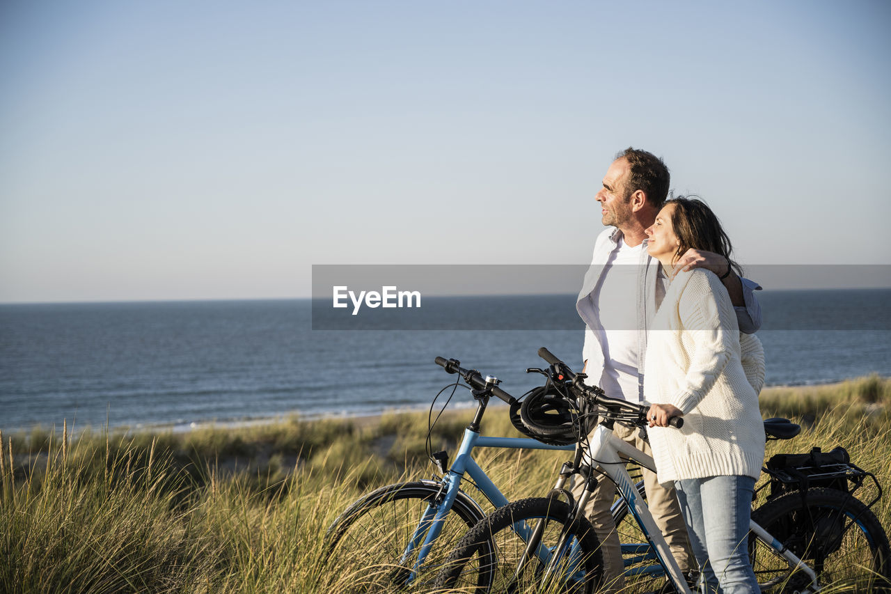 Smiling couple with bicycles looking at view while standing on beach against clear sky during weekend