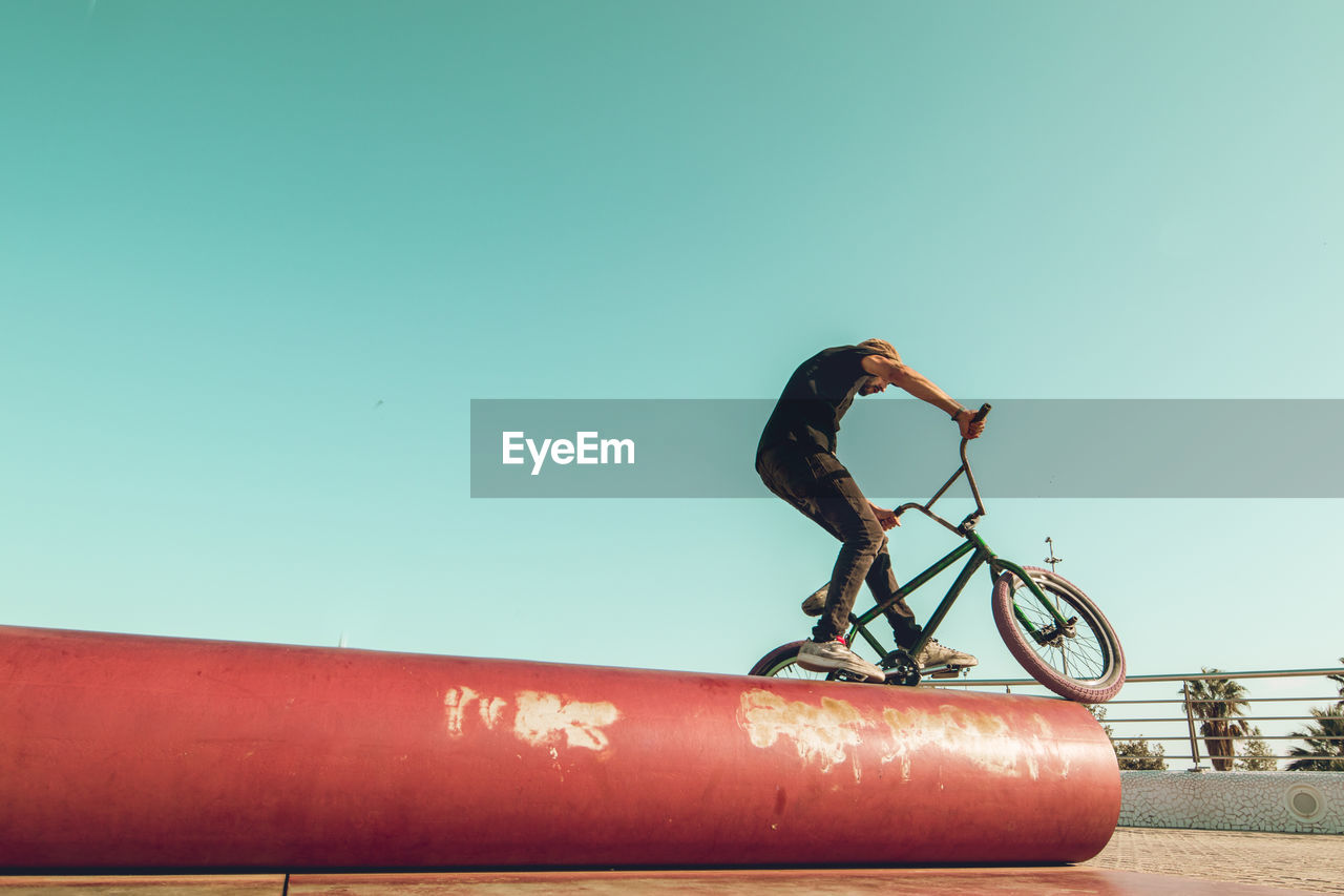 Man riding bicycle on red metallic pipe against sky