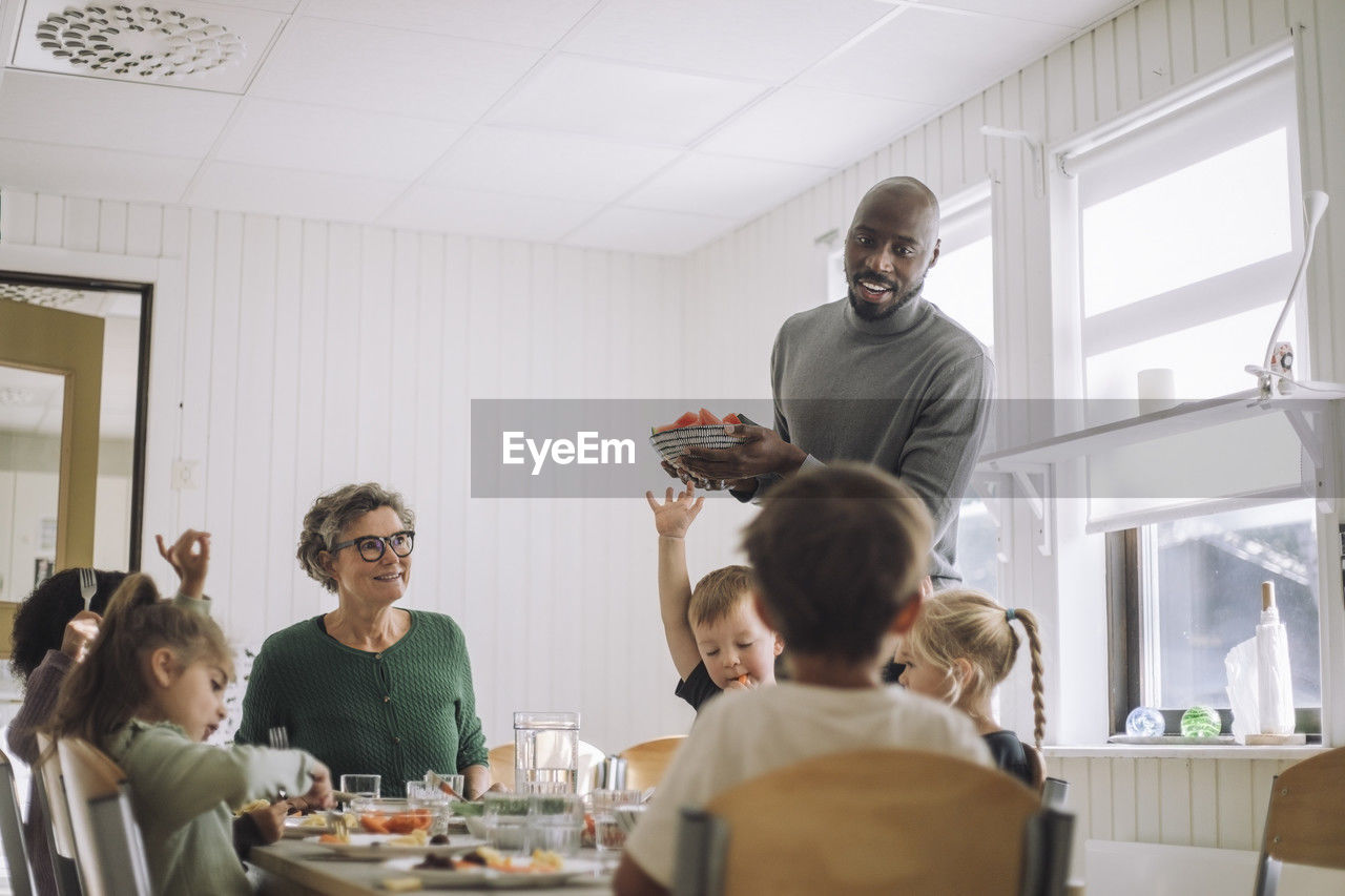 Male teacher serving food to children sitting at dining table during lunch break at kindergarten