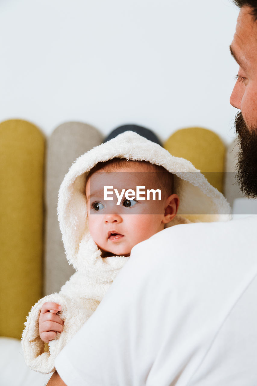 Back view of crop anonymous bearded brutal man embracing carefully infant in bathrobe with interest against blurred interior of light room