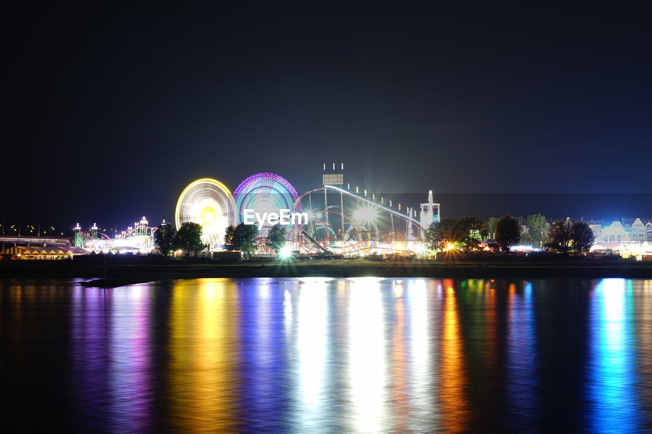 ILLUMINATED FERRIS WHEEL BY RIVER AGAINST SKY AT NIGHT