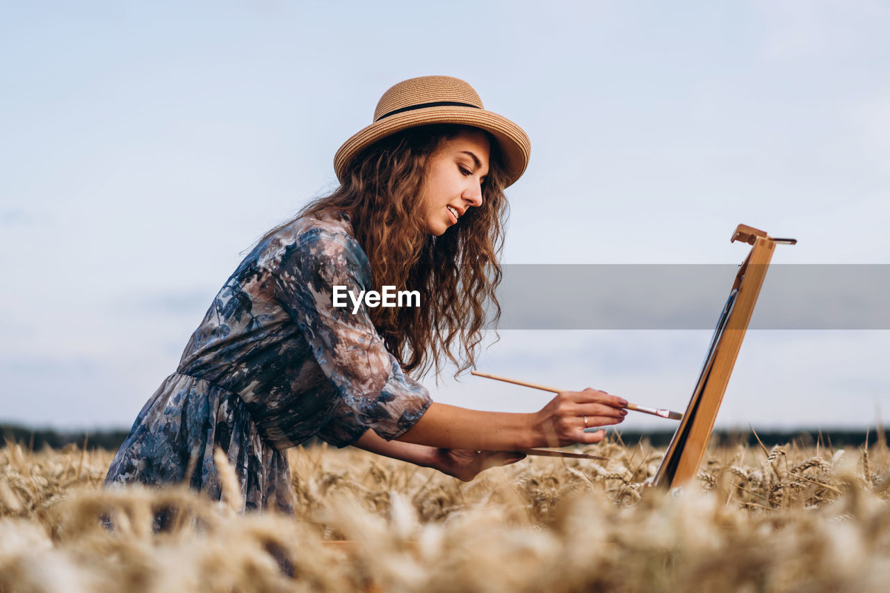 A young woman with curly hair and wearing a hat is painting in nature. 