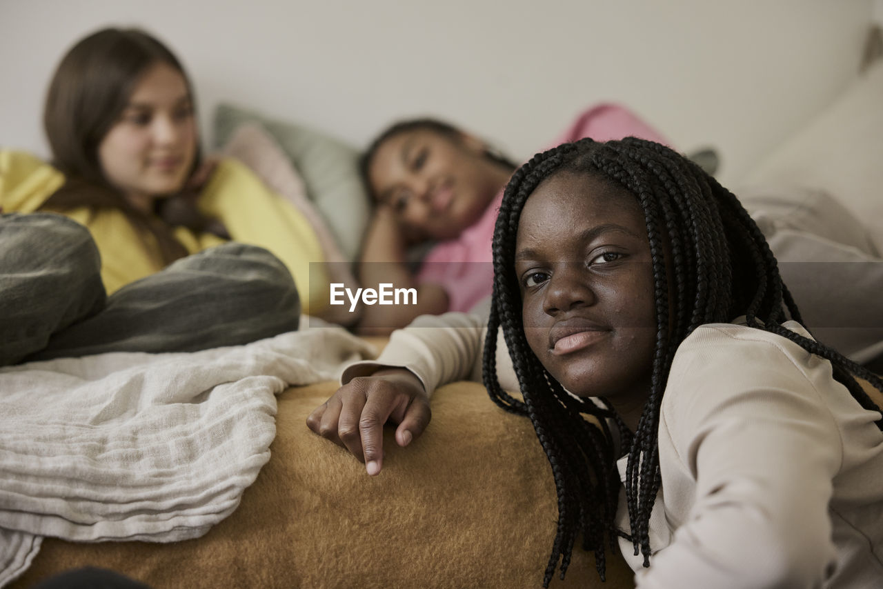 Teenage girl with braided hair by female friends lying on bed in background at home