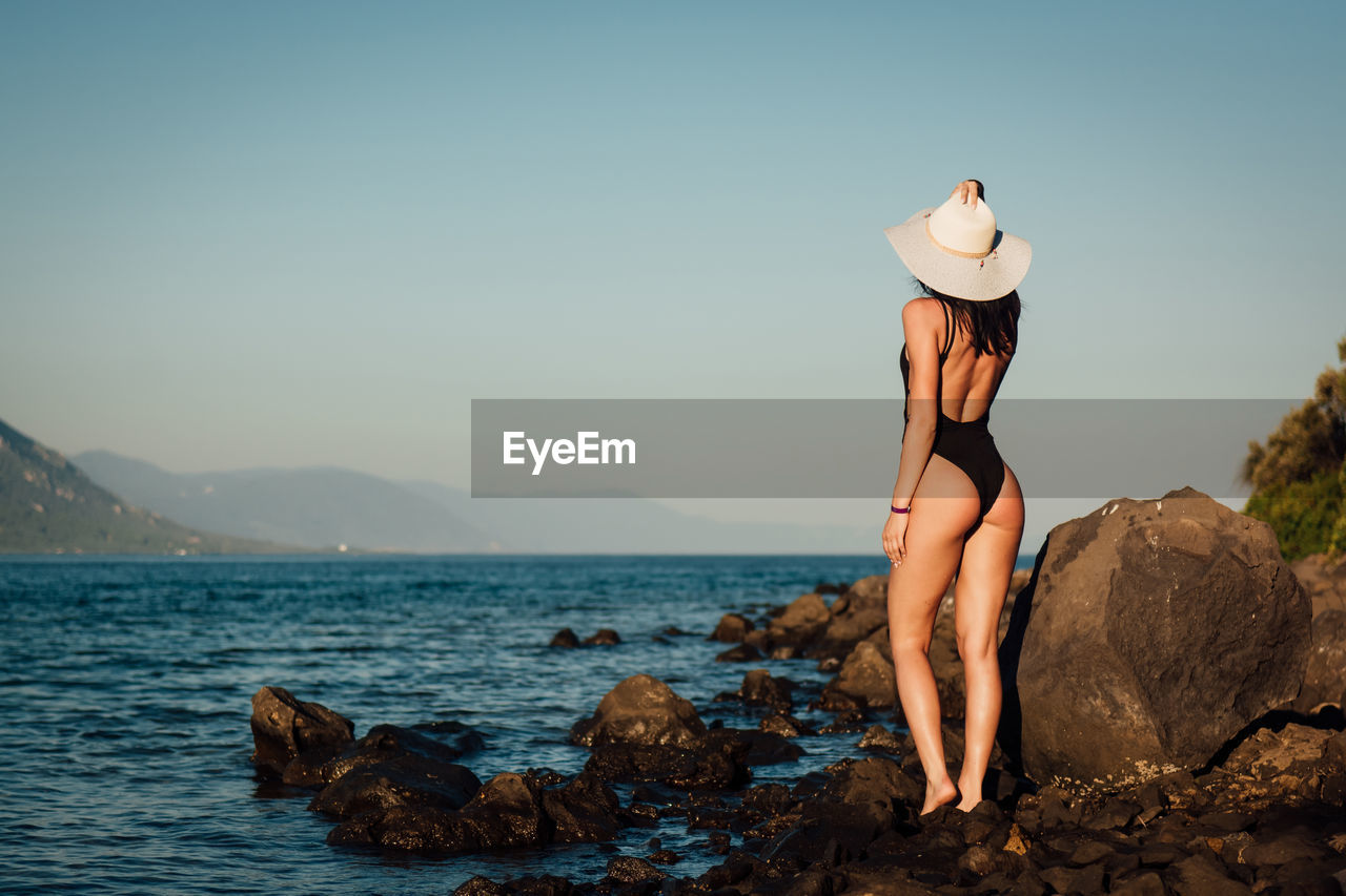 Rear view of sensuous woman standing on rocks at beach against clear sky