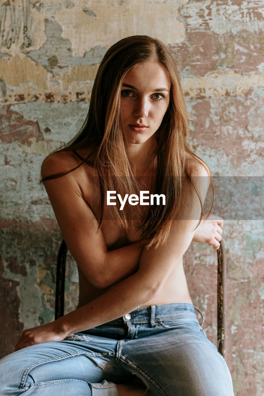 Portrait of shirtless young woman sitting on chair against wall