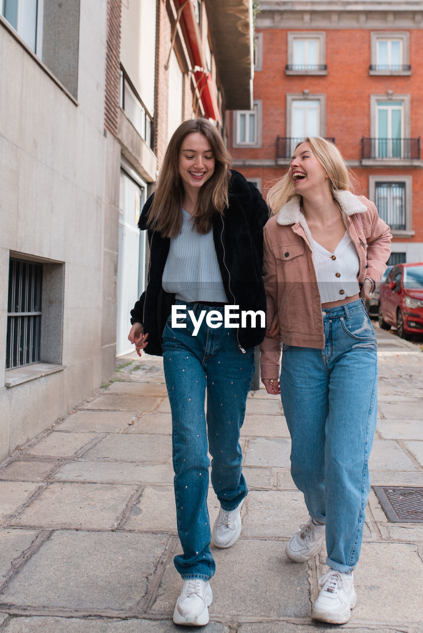 Young stylish female friends walking along street in madrid and laughing while spending weekend together and having fun