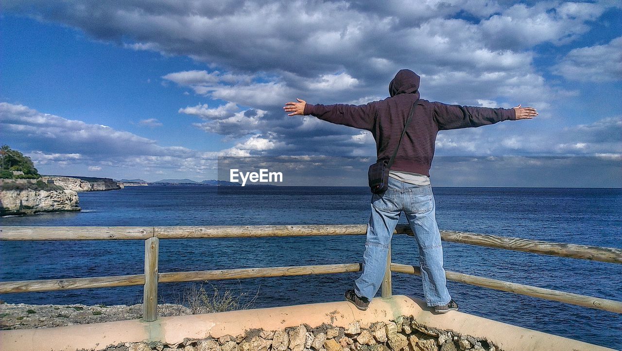 Rear view of man with arms outstretched standing against sea and cloudy sky