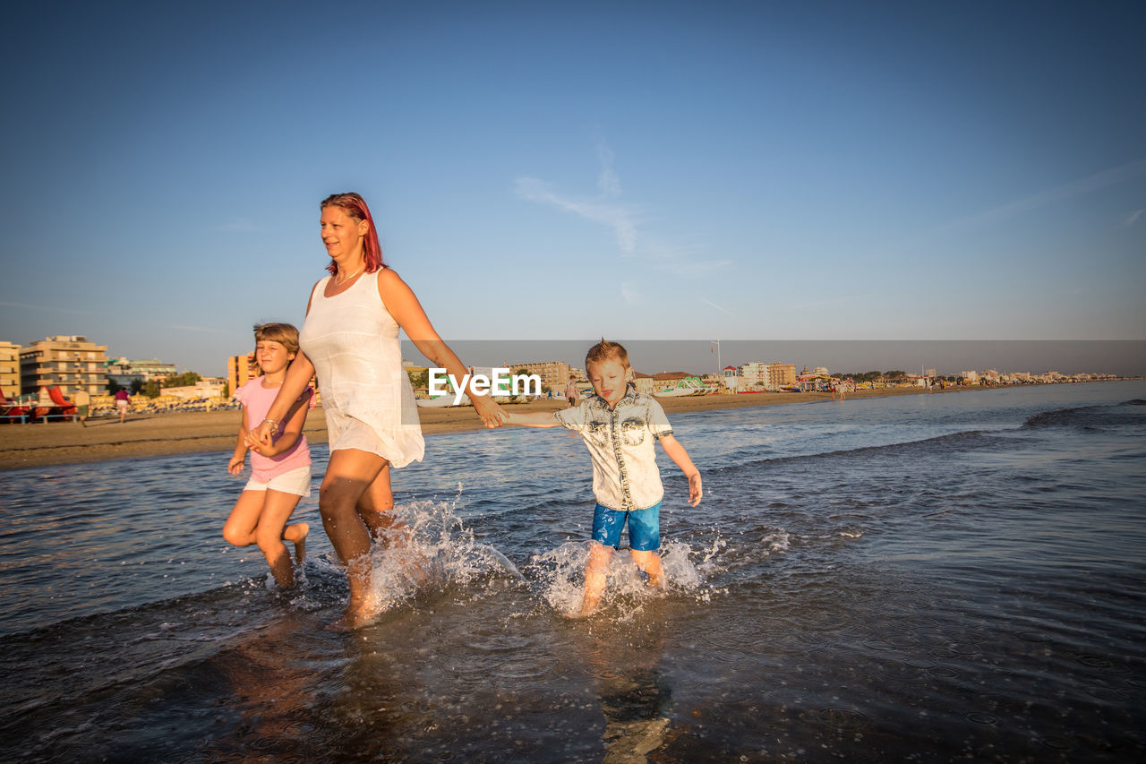 Mother holding children hands while walking in sea on shore at beach during sunset