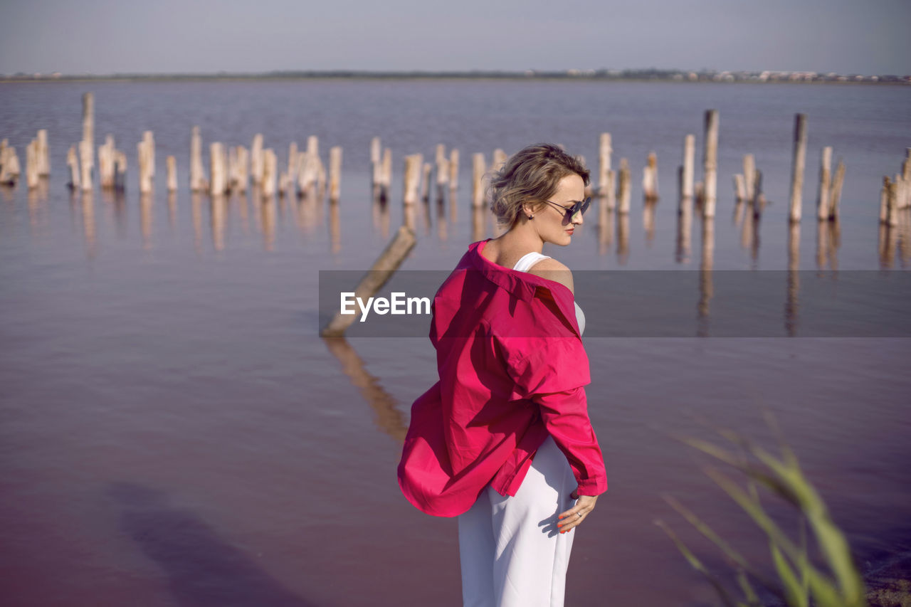 Portrait of young woman in pink and white clothes wearing sunglasses stand on pink lake 