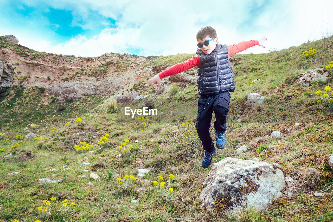 one person, leisure activity, full length, nature, activity, walking, sky, mountain, landscape, hiking, cloud, men, adventure, environment, ridge, land, beauty in nature, child, childhood, lifestyles, wilderness, day, casual clothing, plant, holiday, motion, outdoors, rock, vacation, scenics - nature, trip, travel, adult, grass, vitality, exploration, trail, standing, happiness, sports, recreation, front view, non-urban scene, arm, emotion, limb, backpack, mountain range, clothing, smiling, rural scene, carefree, travel destinations