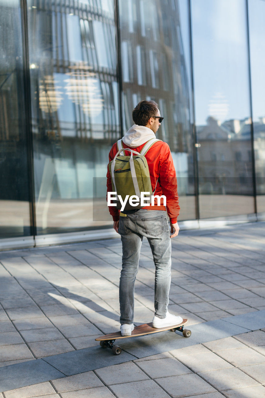 Hipster in red jacket, sunglasses, jeans, white sneakers and with green backpack riding on longboard