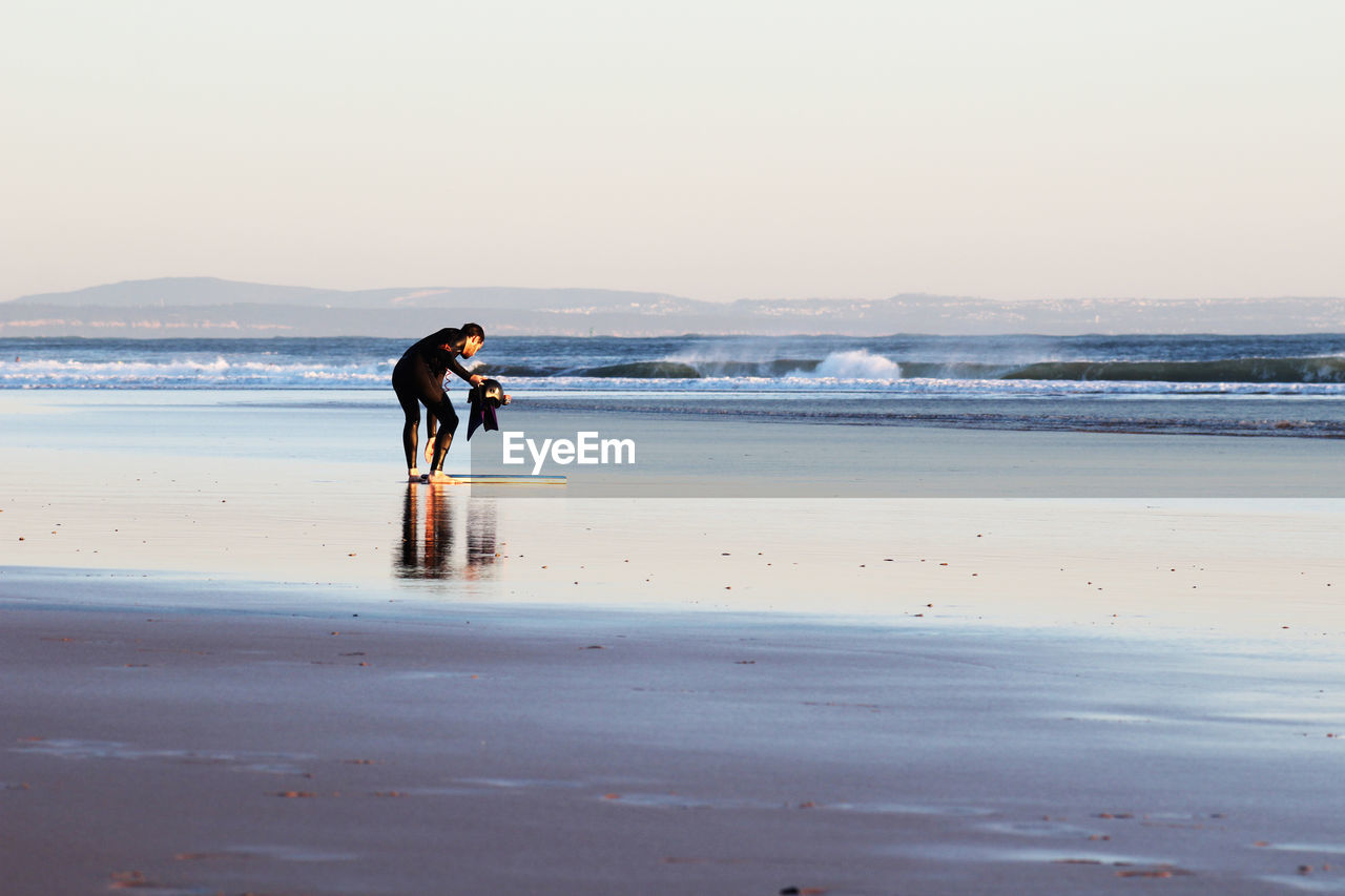 Man holding helmet while bending on sea shore against clear sky