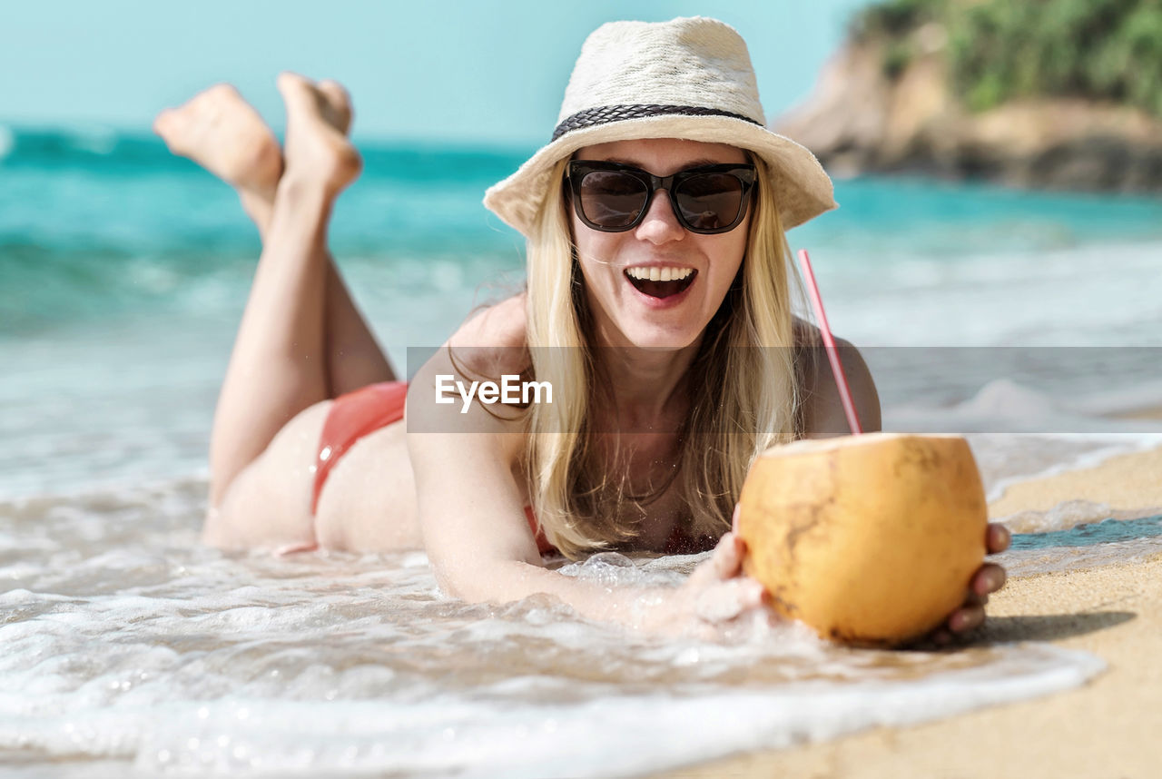 Portrait of smiling young woman in sunglasses,  in summer hat laying on sea beach and drink coconut.