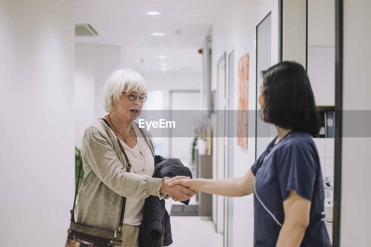Smiling senior woman shaking hands with female doctor standing at doorway in hospital