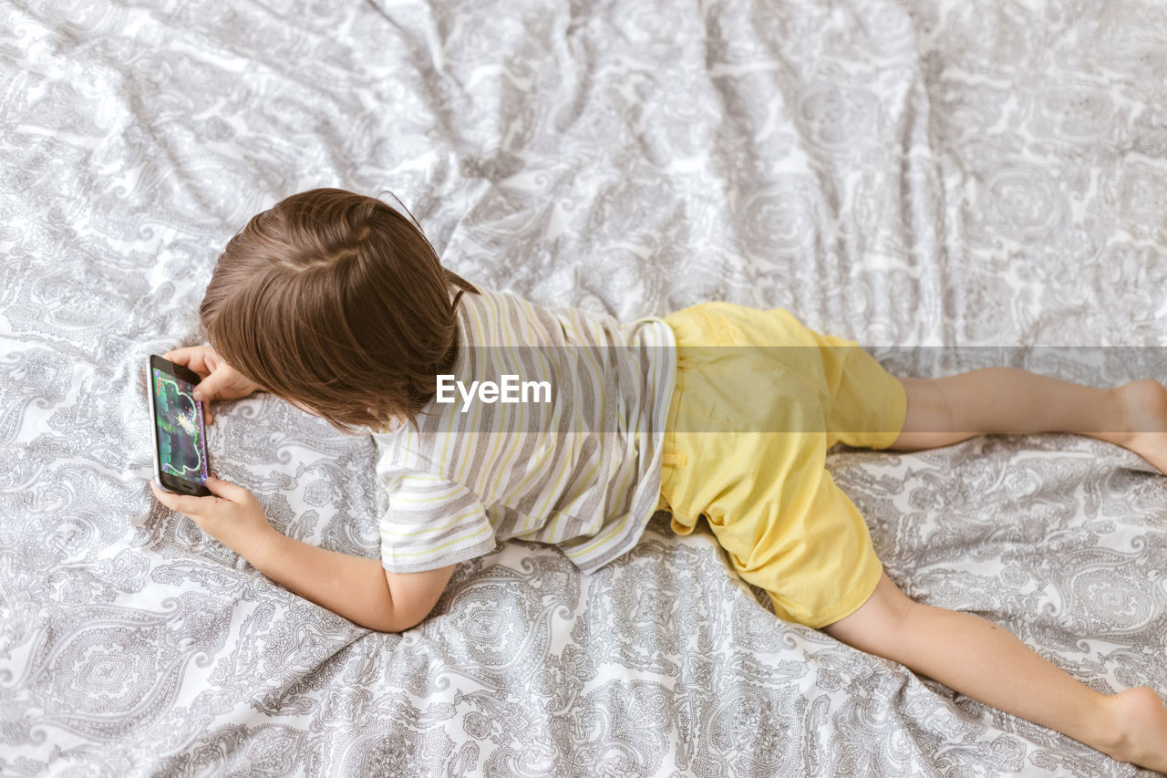 A cute boy, toddler, lies on the bed in the bedroom and plays with the phone, smartphone. 