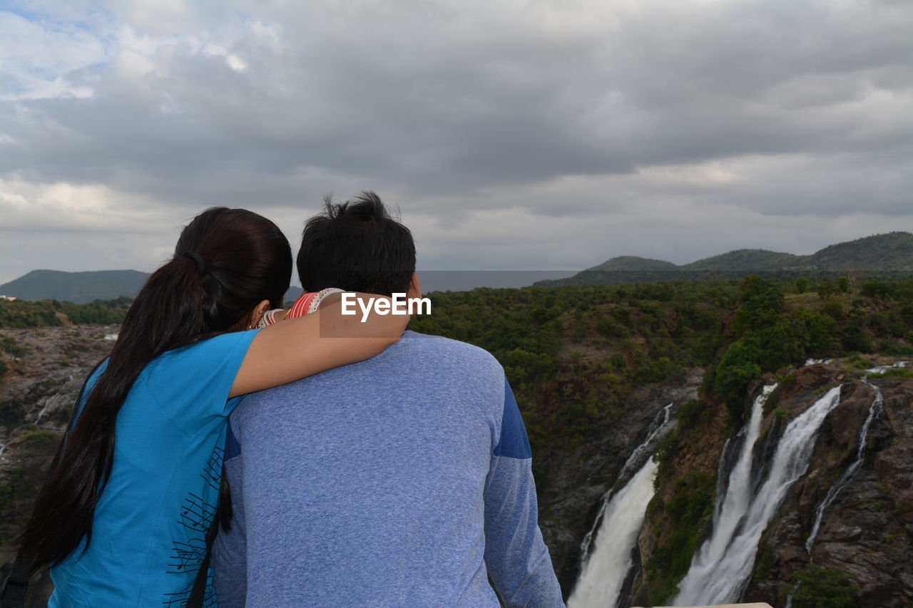 Rear view of young couple looking at waterfall at mountain against cloudy sky