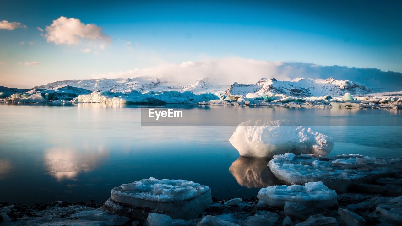 Scenic view of icebergs at lakeshore by snowcapped mountains against sky