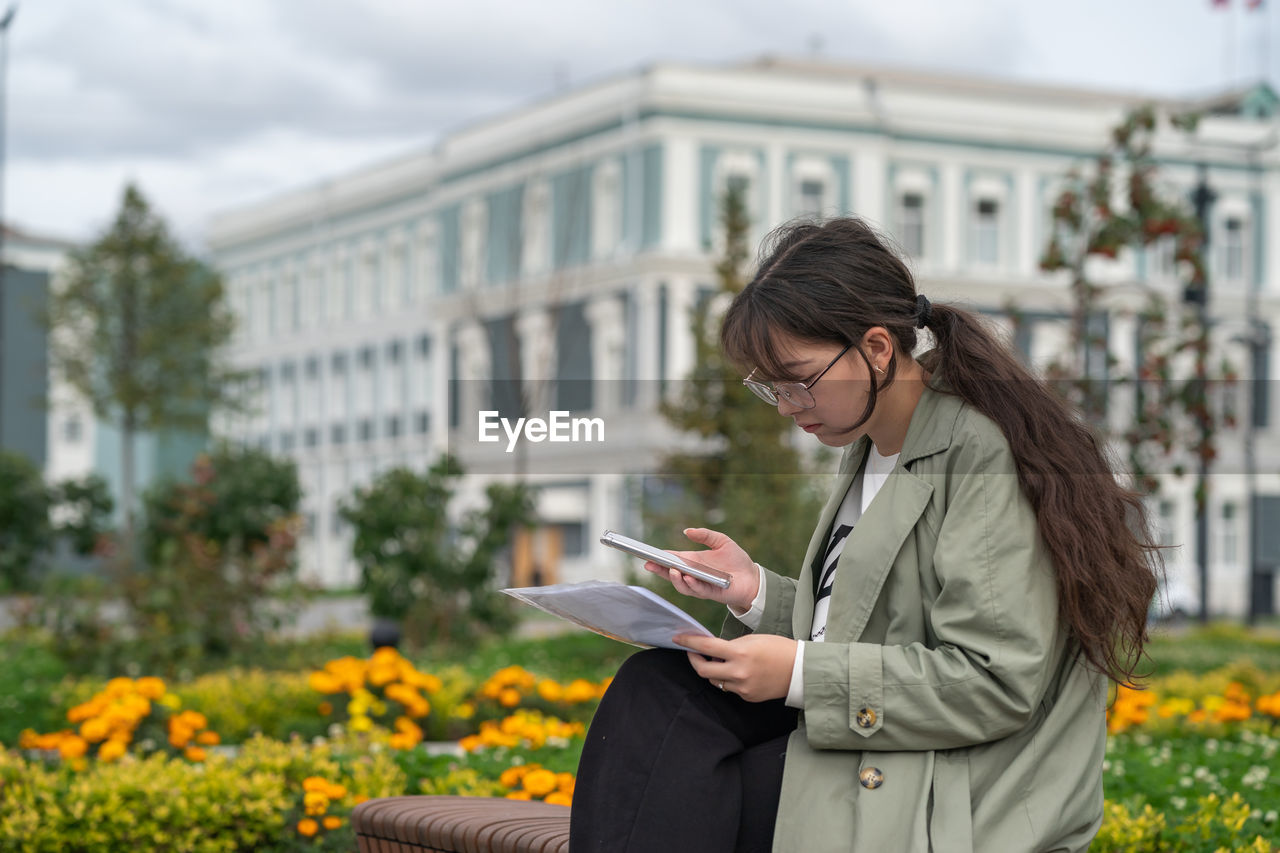 A girl sits on a bench on the street with documents in her hands and using her phone