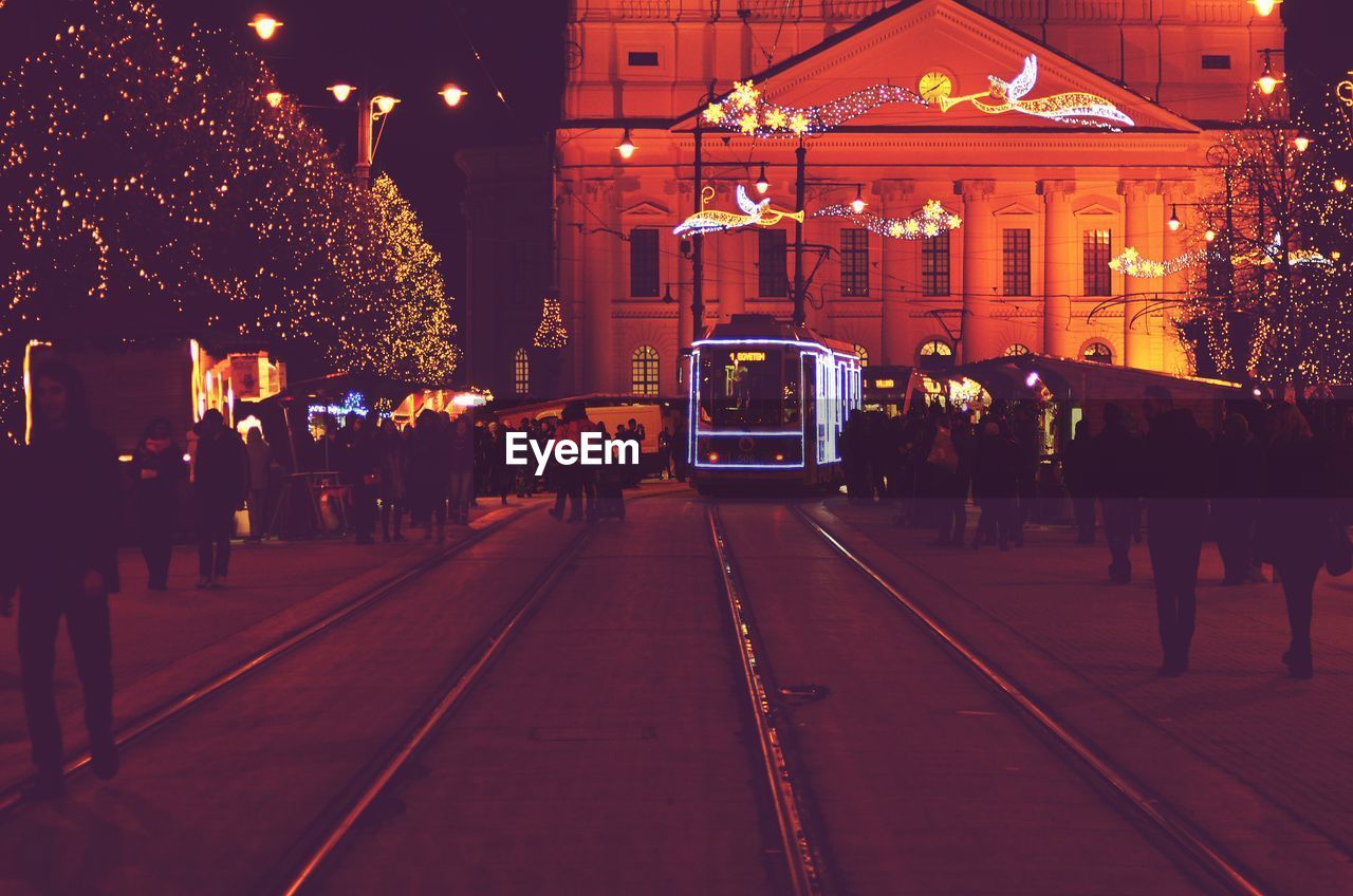 Illuminated tramway moving on street amidst people against decorated buildings