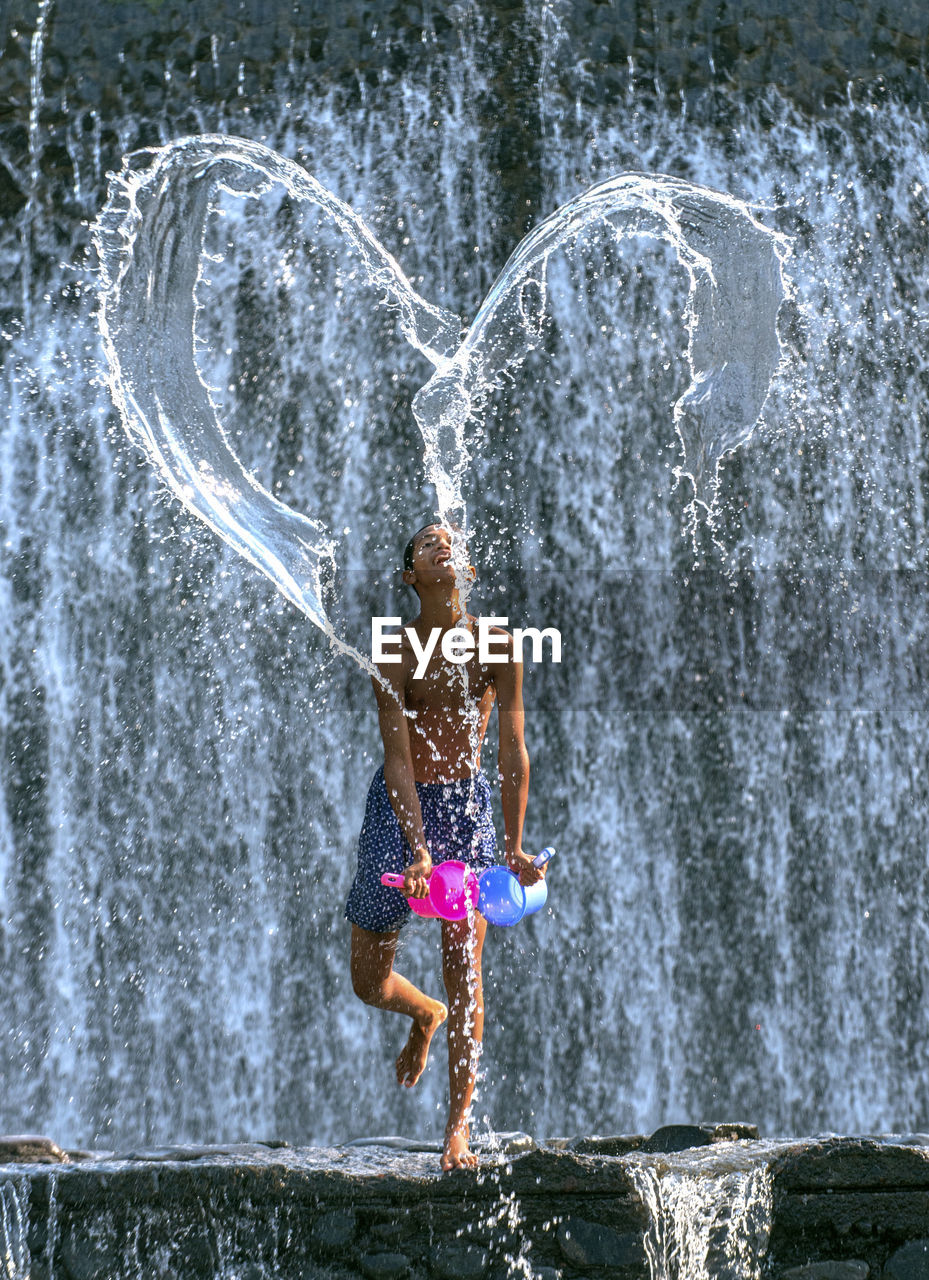 Shirtless man throwing water in heart shape against waterfall