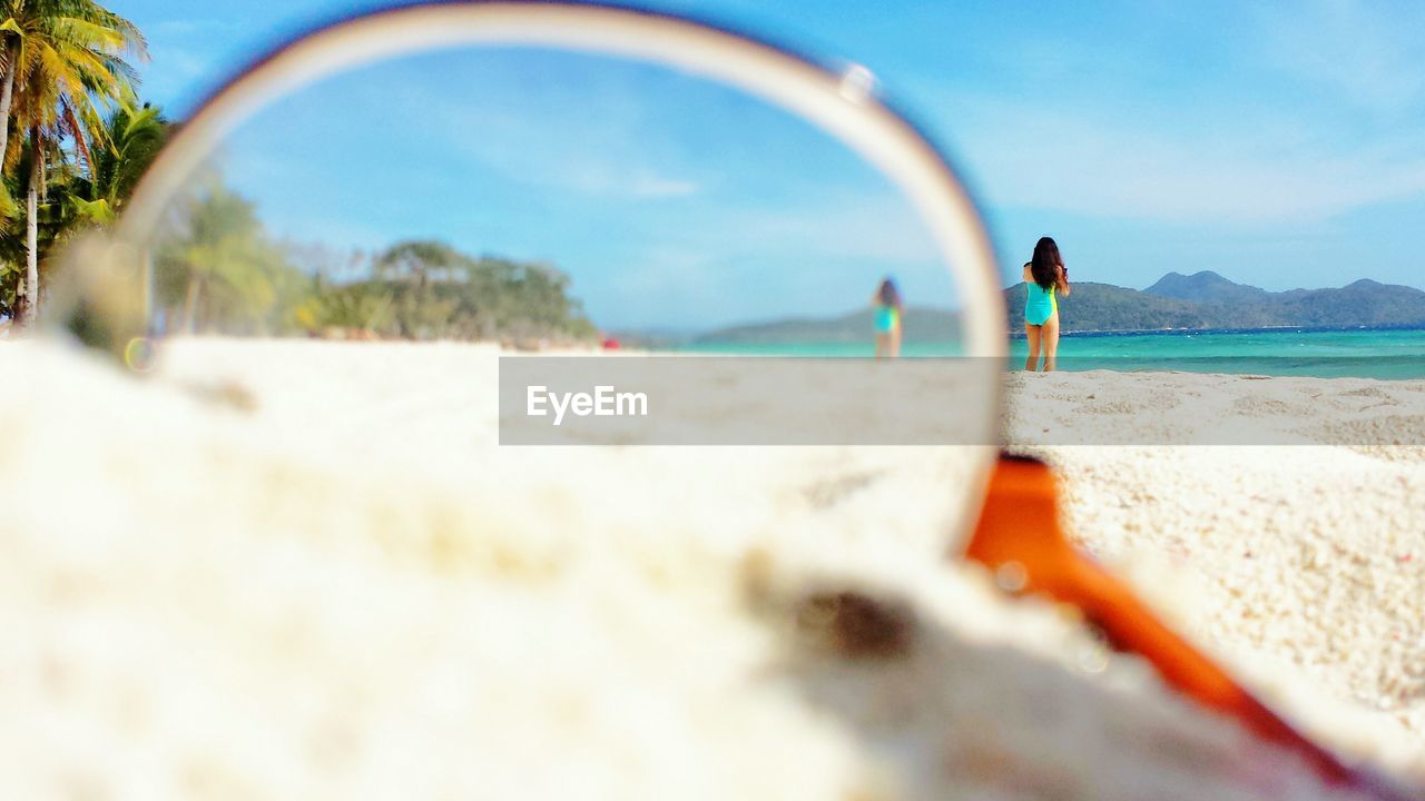 Surface level of eyeglasses on sand with woman in background on beach