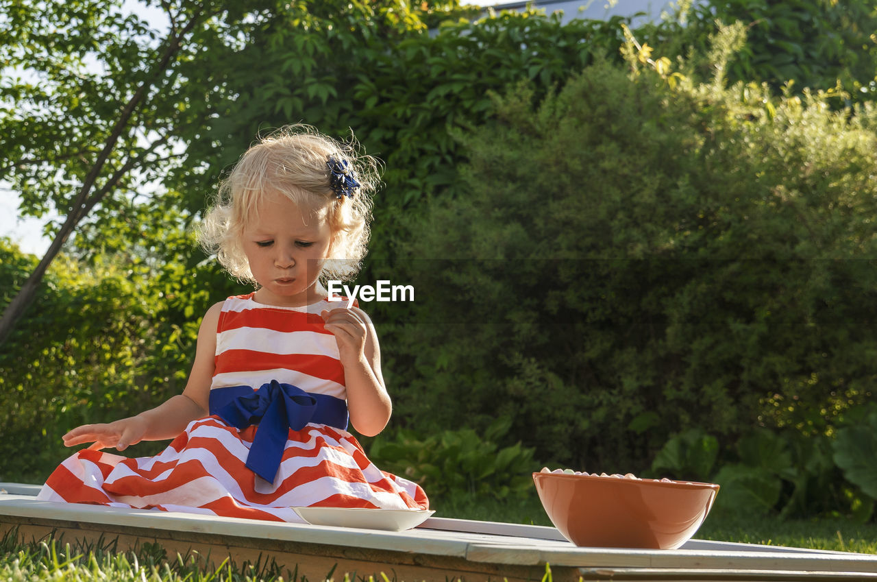 Pretty curly little girl seating in garden and eating marshmallows from the plate.