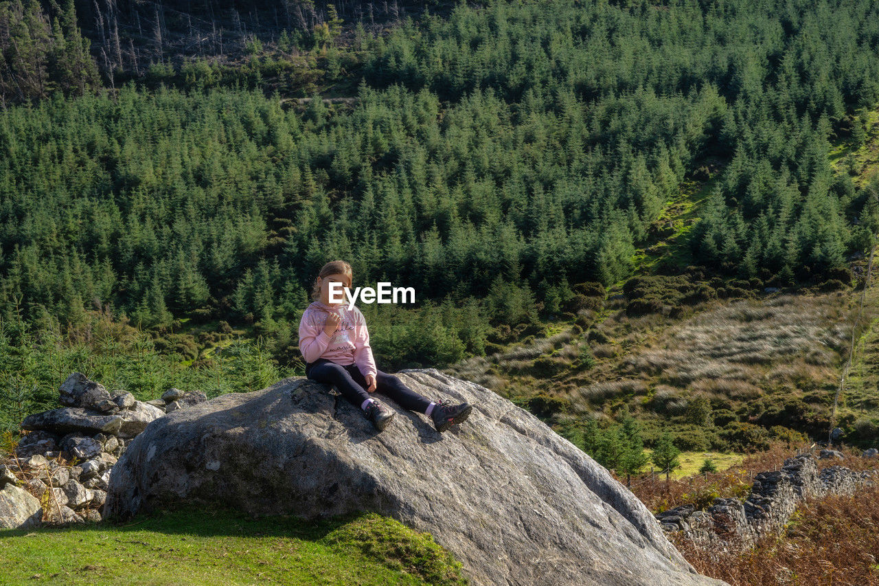 Young girl sitting on rock with forest and valley in background hiking in wicklow mountains, ireland