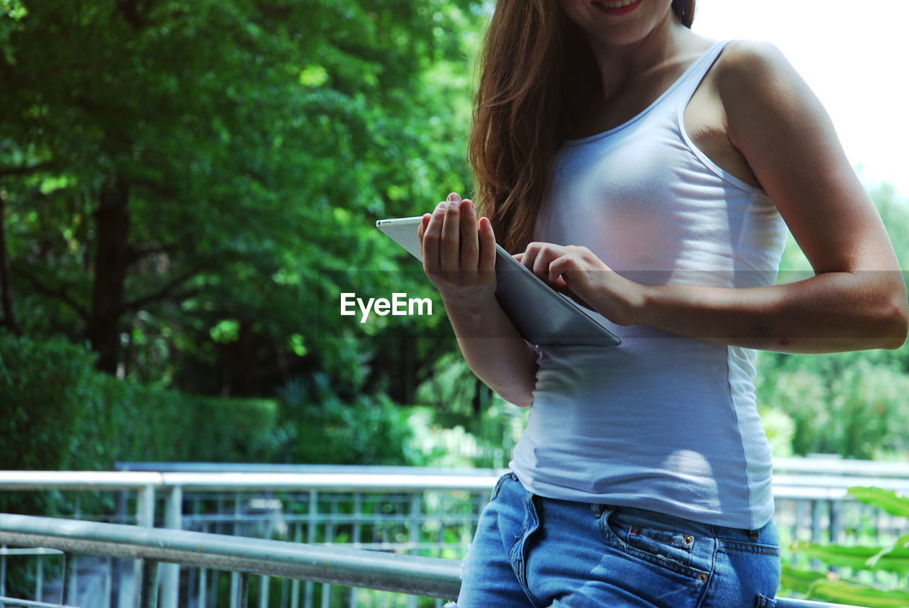 Midsection of woman using digital tablet while leaning on railing against trees