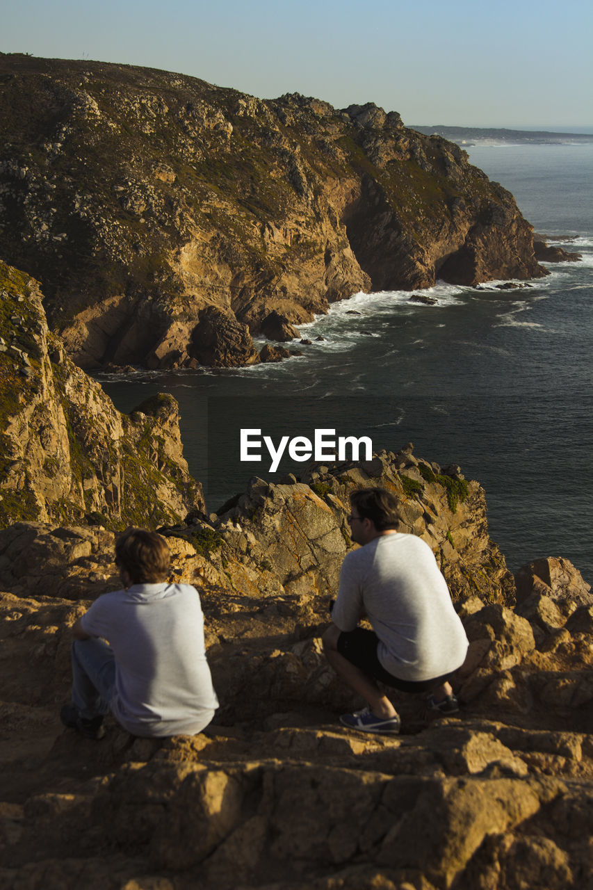 REAR VIEW OF PEOPLE SITTING ON ROCKS AT SEA SHORE