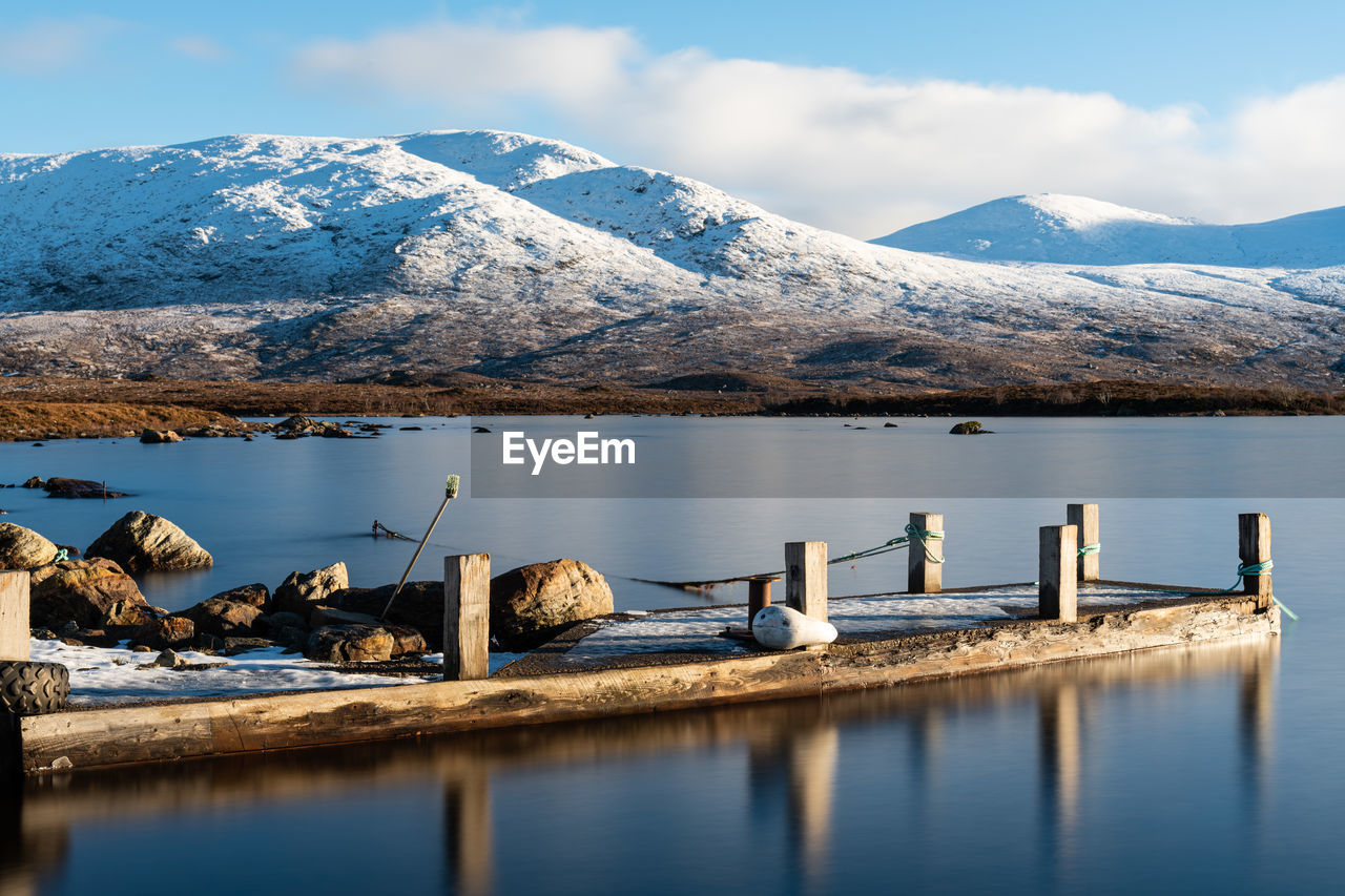 WOODEN POSTS IN LAKE AGAINST SNOWCAPPED MOUNTAINS