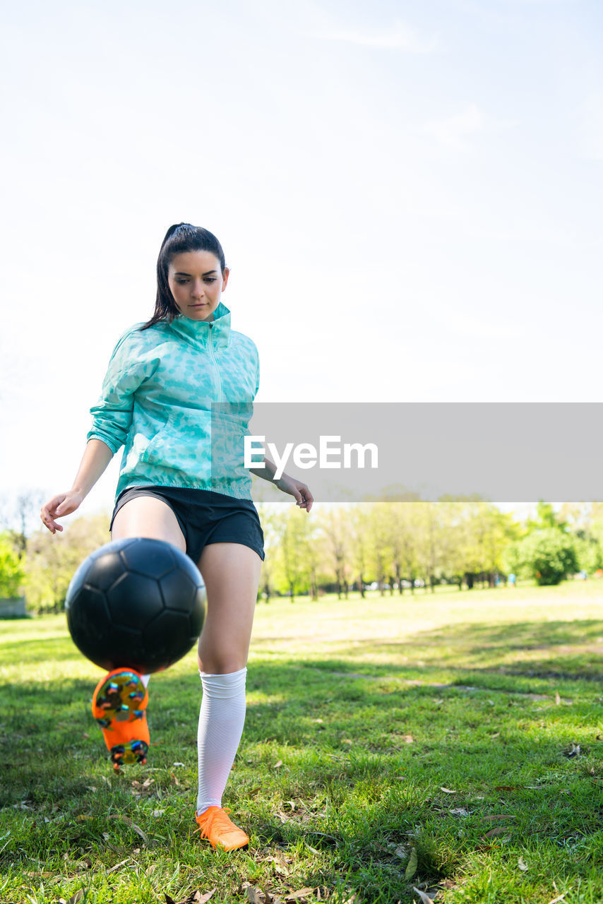 Full length of young woman playing soccer on field against sky