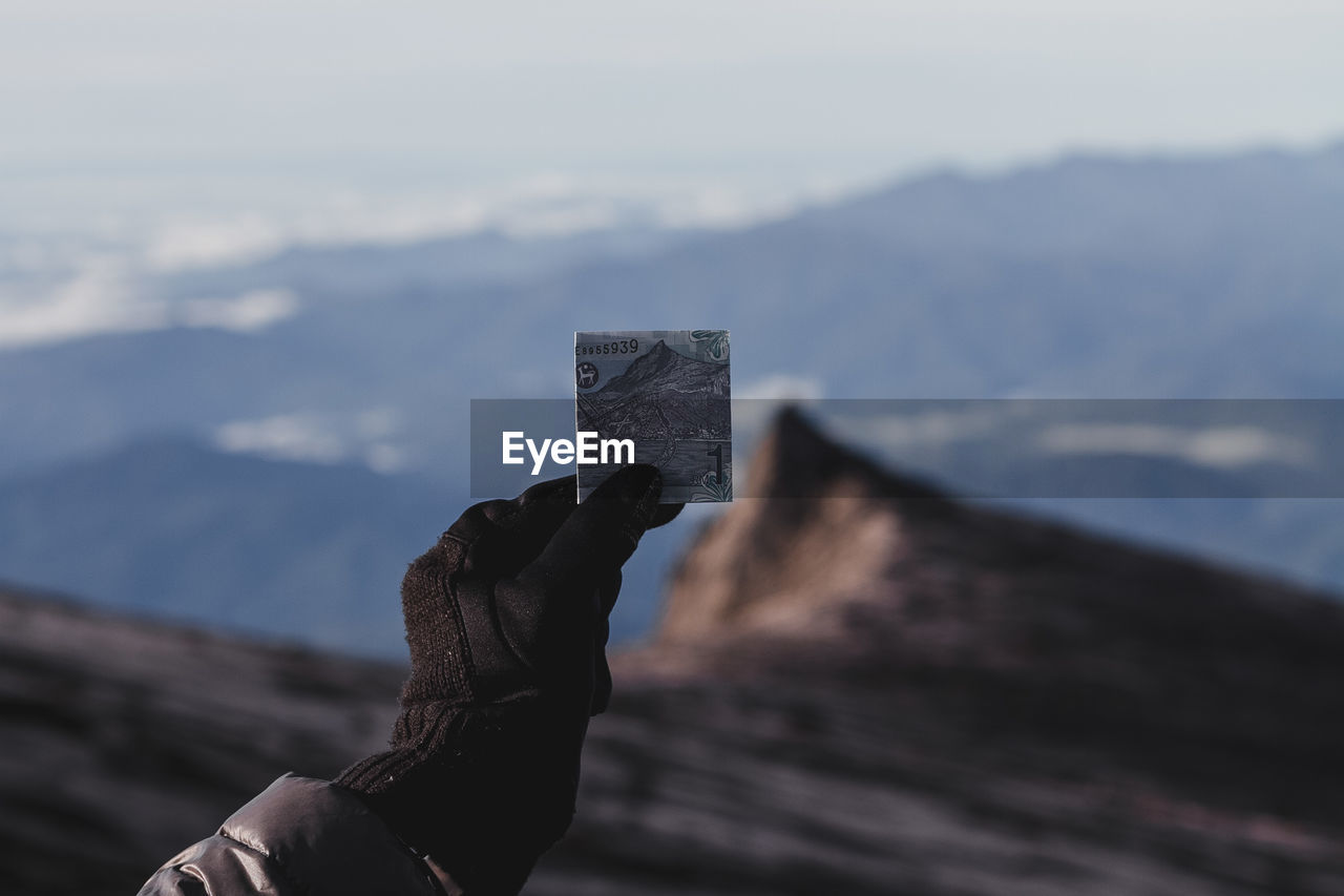 Cropped hand of person holding paper currency on mountains against sky