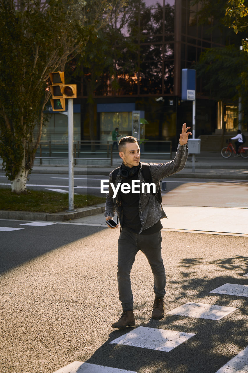 Young male crossing road in city and waving hand while looking away and catching taxi