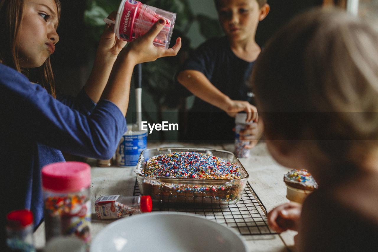 Siblings decorating cake with overabundance of colorful sprinkles