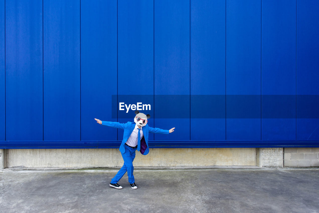 Man wearing vibrant blue suit and panda mask standing outdoors with raised arms