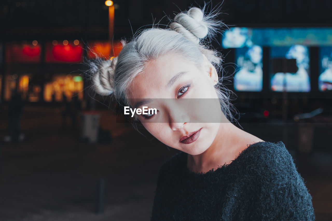 Informal asian female adolescent with dyed blond hair and bright makeup looking at camera while standing on night city street