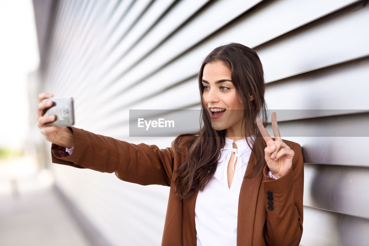 Young businesswoman taking selfie while gesturing peace sign by blinds