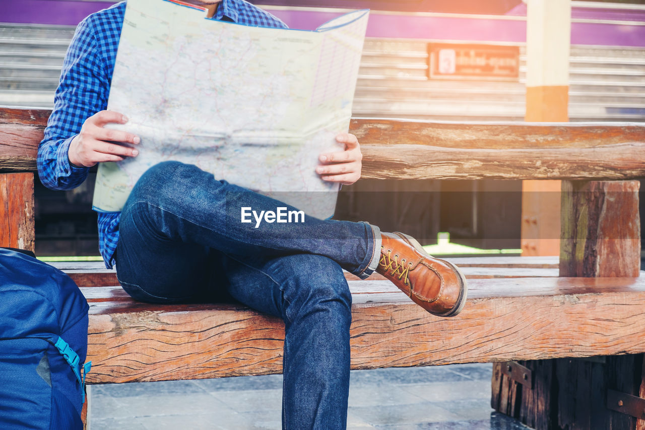 Low section of man reading map while sitting on bench at railroad station