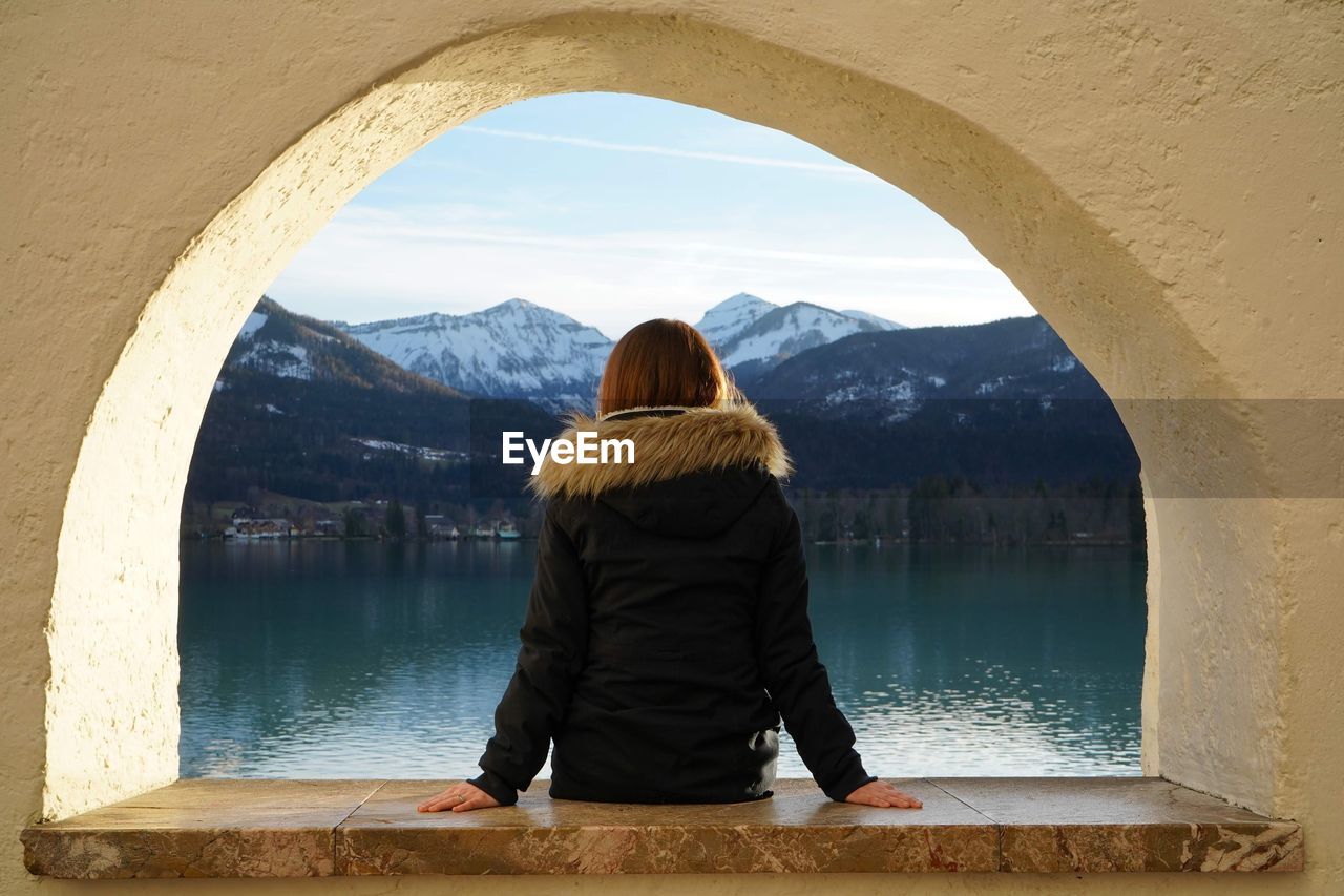 Rear view of woman sitting on arched window over lake against mountains and sky
