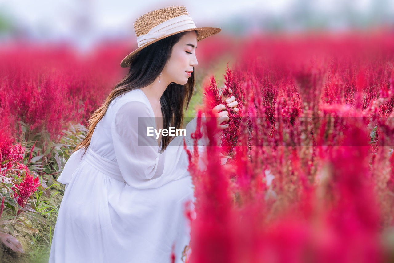 women, plant, flower, flowering plant, adult, one person, beauty in nature, field, young adult, nature, clothing, red, hat, land, landscape, rural scene, fashion, pink, freshness, long hair, fashion accessory, smiling, female, summer, dress, lifestyles, environment, happiness, spring, relaxation, agriculture, sky, hairstyle, emotion, tranquility, selective focus, tranquil scene, springtime, growth, sun hat, outdoors, side view, meadow, looking, person, portrait, leisure activity, idyllic, plain, positive emotion, standing, white, farm, grass, cheerful, blossom, contemplation, elegance, non-urban scene, crop, multi colored