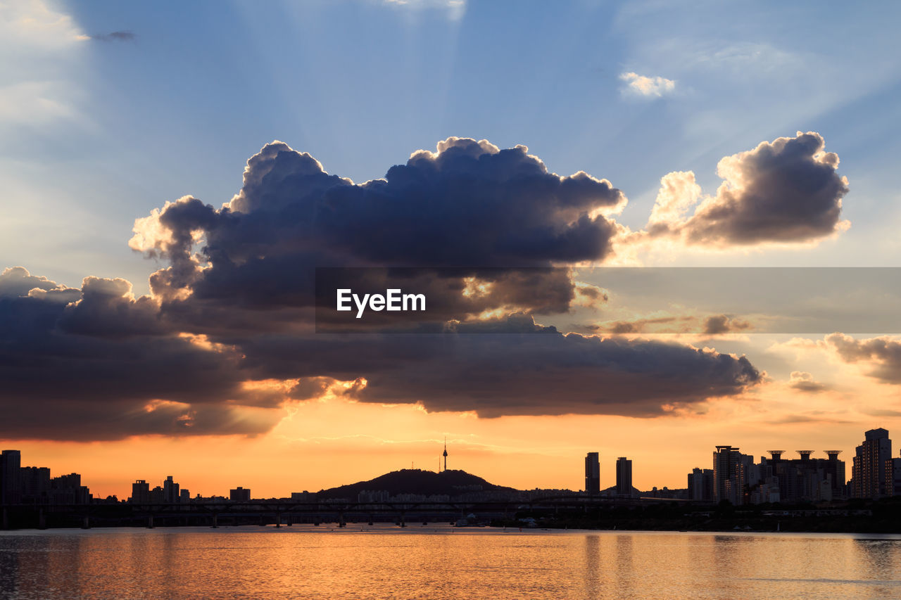 SILHOUETTE BUILDINGS BY RIVER AGAINST SKY DURING SUNSET