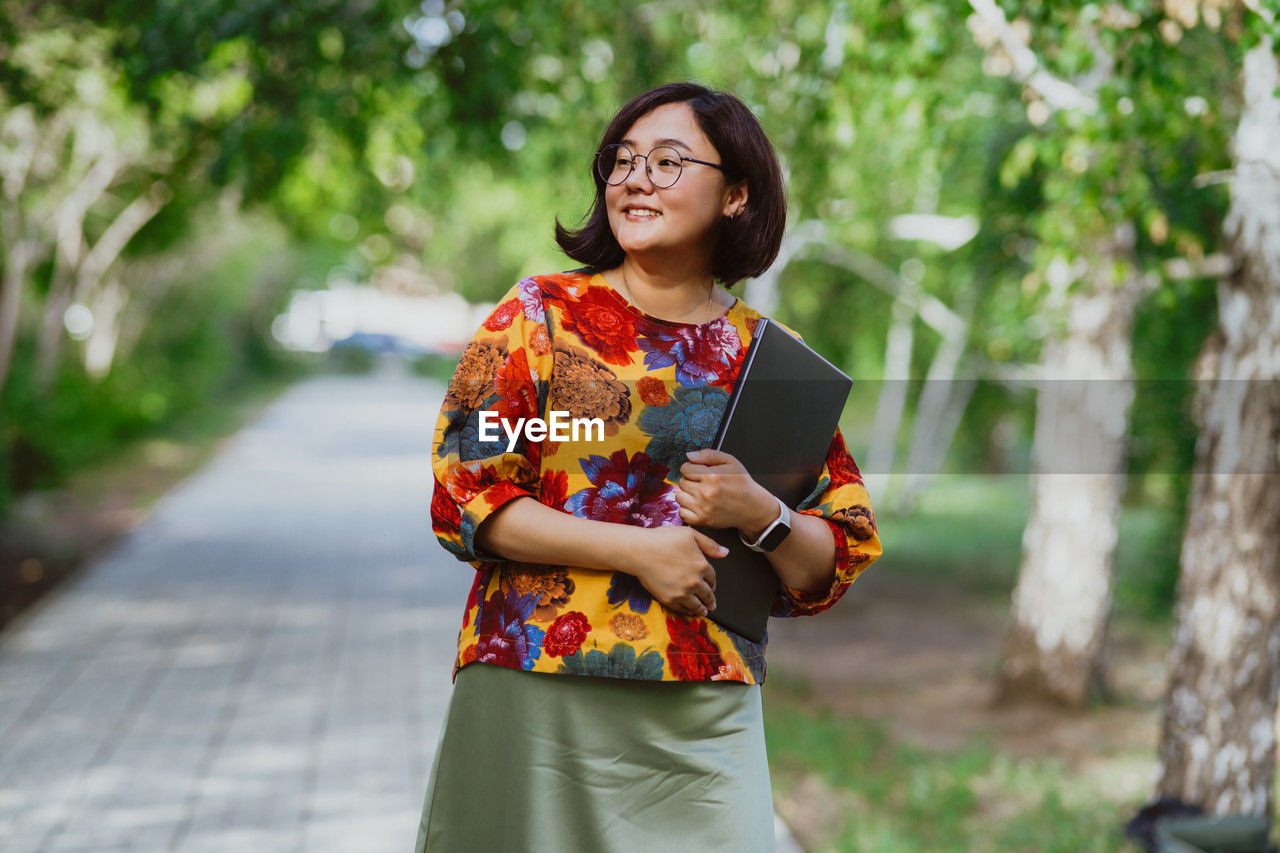 A smiling asian woman with glasses holding a laptop in a sunny park.