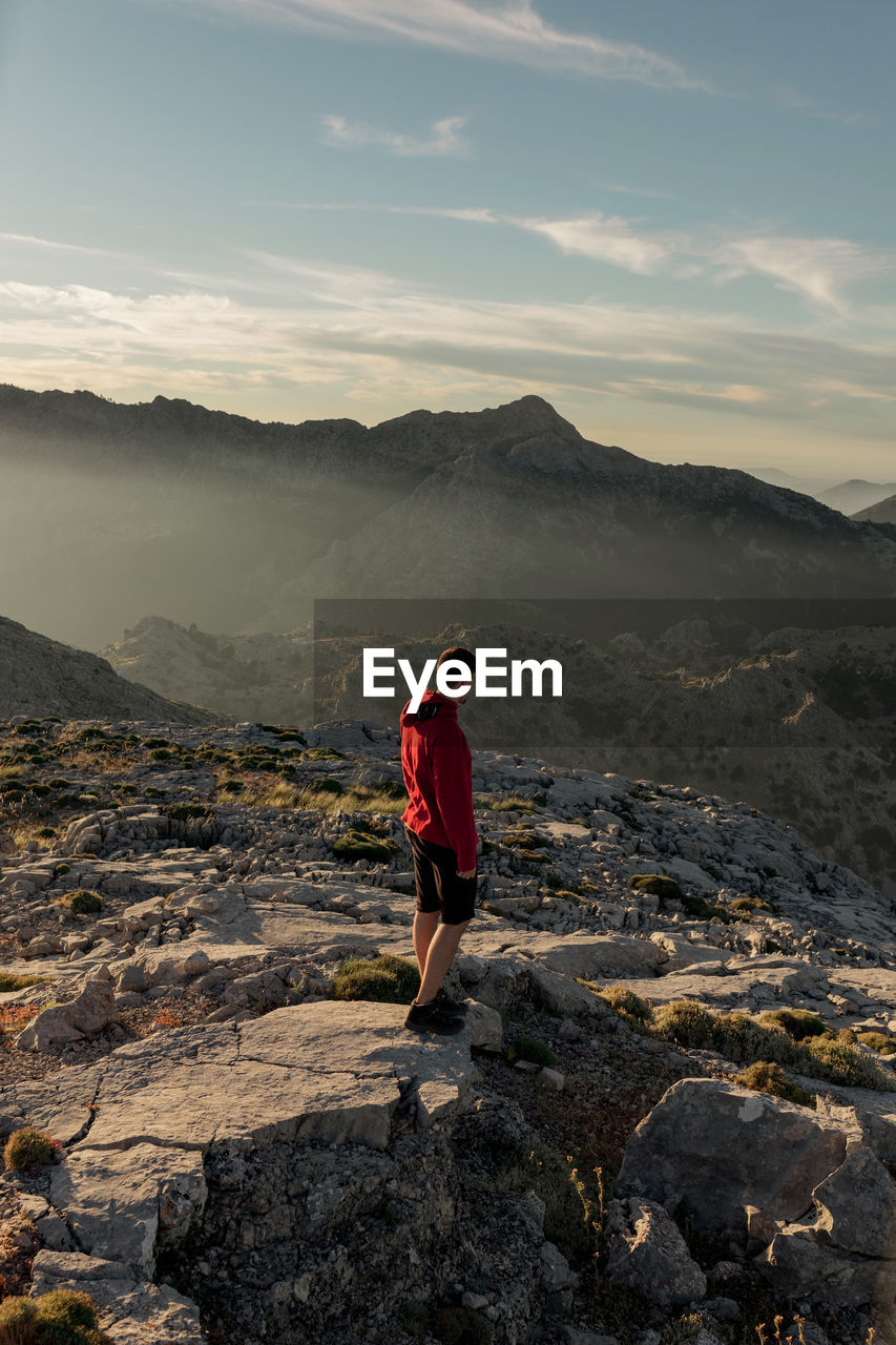 Back view anonymous male hiker in casual wear resting on rocky mountain summit and admiring scenic views over rough highlands in seville spain